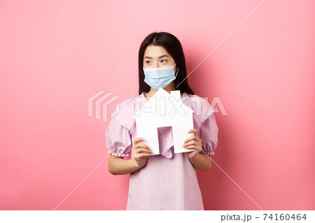 Real estate and pandemic concept. Young asian woman in medical mask dreaming of home, showing paper house cutout and look aside, standing against pink background 74160464
