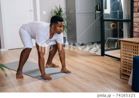 African-American man practicing burpee exercise at home, doing push-ups and jumping on yoga mat. 74165325