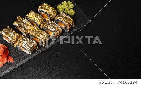 Custom sushi roll with nori, cuttlefish ink, fresh salmon, cream cheese, pepper pumpkin, eel, unagi sauce and edible gold leaf with red ginger, wasabi on a square plate on black table and background. 74165384