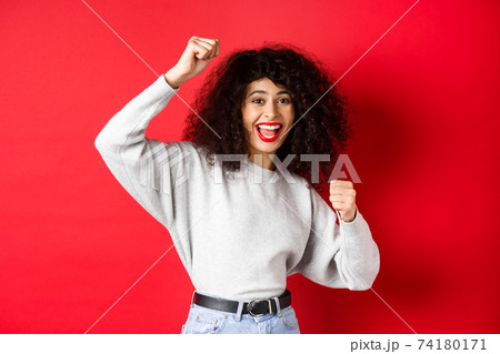 Cheerful young woman with curly hair, raising hand up and celebrating win, achieve goal or success, standing on red background 74180171