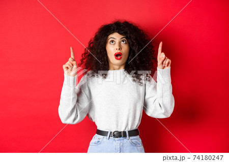 Impressed european female model in casual clothes, checking out promo with dropped jaw, looking and pointing fingers up, showing logo, standing on red background 74180247