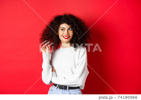 Modern woman in sweatshirt, playing with curly hair and looking aside at empty space, standing on red background Modern woman in sweatshirt, playing with curly hair and looking aside at empty space, standing on red background 74180966