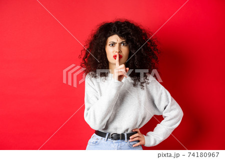 Angry young woman with curly hair and red lips, frowning and shushing, tell to be quiet, make taboo gesture to keep mouth shut, standing on red background 74180967