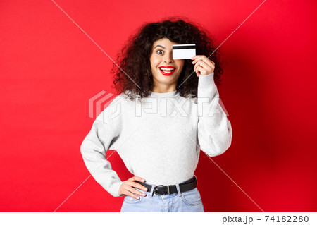 Stylish girl with curly hair showing plastic credit card over eye and smiling, standing against red background Stylish girl with curly hair showing plastic credit card over eye and smiling, standing against red background 74182280