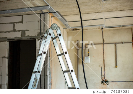 Aluminium ladder and electrical cables laid in protective corrugation installed on the ceiling and wall in a room under construction works. Aluminium ladder and electrical cables laid in protective corrugation installed on the ceiling and wall in a room under construction works. 74187085