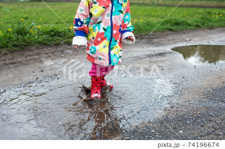 A little girl in a bright jacket and red boots jumps merrily through puddles. 74196674