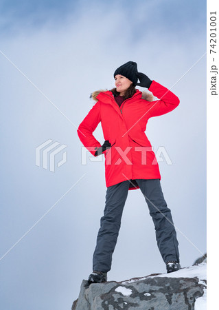Young woman standing on top of mountain peak on background of sky with clouds. Smiling woman dressed in red jacket, sports pants, trekking boots Young woman standing on top of mountain peak on background of sky with clouds. Smiling woman dressed in red jacket, sports pants, trekking boots 74201001