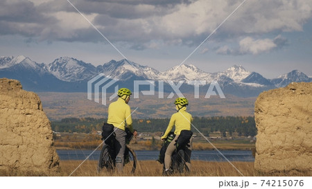 The man and woman travel on mixed terrain cycle touring with bikepacking. The two people journey with bicycle bags. Mountain snow capped, stone arch. The man and woman travel on mixed terrain cycle touring with bikepacking. The two people journey with bicycle bags. Mountain snow capped, stone arch. 74215076