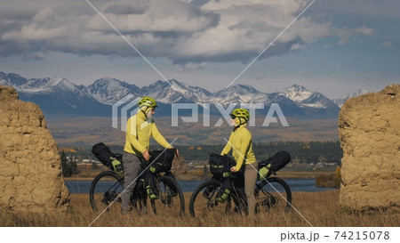 The man and woman travel on mixed terrain cycle touring with bikepacking. The two people journey with bicycle bags. Mountain snow capped, stone arch. The man and woman travel on mixed terrain cycle touring with bikepacking. The two people journey with bicycle bags. Mountain snow capped, stone arch. 74215078