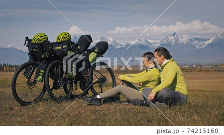 The man and woman travel on mixed terrain cycle touring with bikepacking. The two people journey with bicycle bags. Mountain snow capped. 74215160
