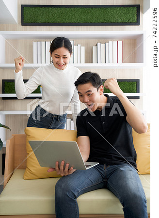 Young lovers spend time together on holidays in the living room. Both raised their fist up and smiled happily at the good news they received in e-mail while the man hold laptop computer. Young lovers spend time together on holidays in the living room. Both raised their fist up and smiled happily at the good news they received in e-mail while the man hold laptop computer. 74222596