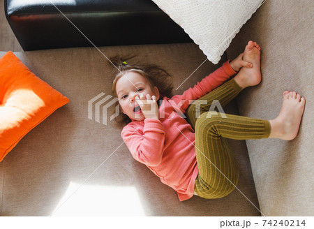 a little girl in a pink sweatshirt is lying on the sofa among the pillows with her legs up and picking her nose. Home games. a little girl in a pink sweatshirt is lying on the sofa among the pillows with her legs up and picking her nose. Home games. 74240214