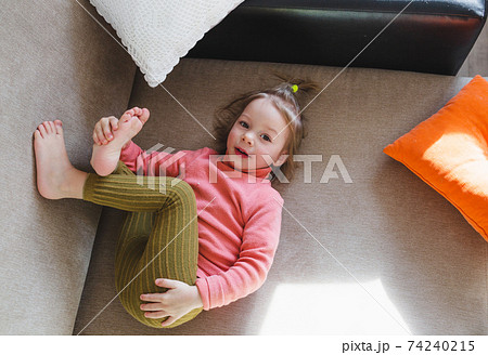 a little girl in a pink sweatshirt is lying on the sofa among the pillows with her legs up. Home games. a little girl in a pink sweatshirt is lying on the sofa among the pillows with her legs up. Home games. 74240215