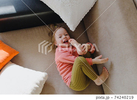 a little girl in a pink sweatshirt is lying on the sofa among the pillows with her legs up. Home games. a little girl in a pink sweatshirt is lying on the sofa among the pillows with her legs up. Home games. 74240219