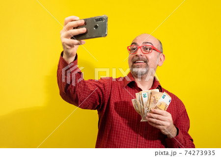 Middle-aged Caucasian man holding a bunch of 50 euro bills in one hand and taking a selfie with the other. He is happily surprised. Concept of winning, luck and wealth. Yellow background. Middle-aged Caucasian man holding a bunch of 50 euro bills in one hand and taking a selfie with the other. He is happily surprised. Concept of winning, luck and wealth. Yellow background. 74273935