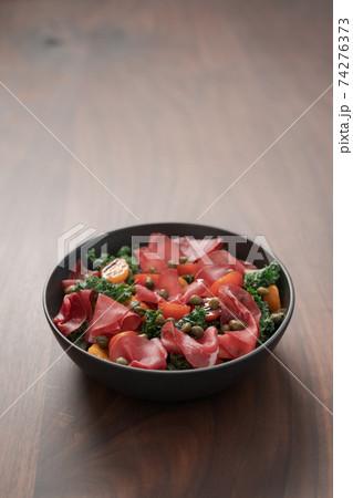 Salad with kale, bresaola and cherry tomatoes decorated with capers in black bowl on walnut wood table 74276373