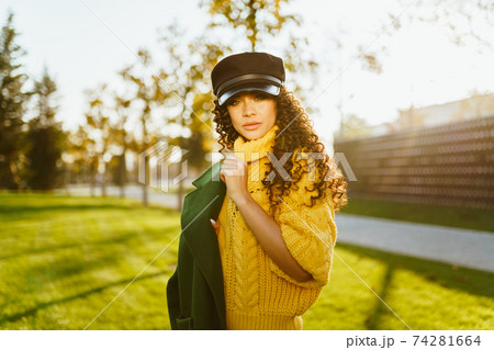 In the autumn park, a girl in a leather cap and a bright yellow sweater slung over her shoulder a green jacket with her hair with dark curls scattered over her shoulders In the autumn park, a girl in a leather cap and a bright yellow sweater slung over her shoulder a green jacket with her hair with dark curls scattered over her shoulders 74281664