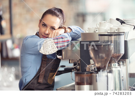 Bored waitress leans against the coffee machine with no job to do, holding face mask in her hand 74283203