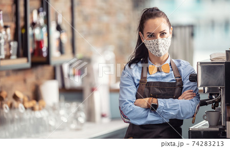 Female employee in a cafe stands unhappy with arms crossed wearing face mask 74283213