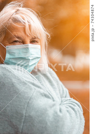 Caucasian blonde woman 50 years old in a cozy warm sweater and a protective medical mask looking at the camera in an autumn park 74304874