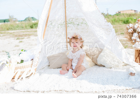 A little cute girl with red curly hair in summer, on the seashore in an airy lace bodysuit and a floral headband in a lace tent, devoried with marine details, shells and cotton. 1st BIRTHDAY.  74307366