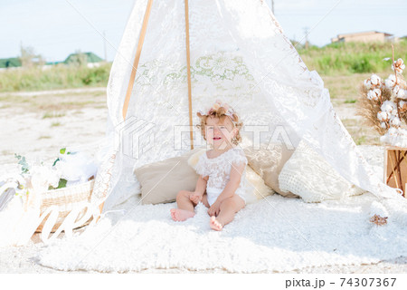 A little cute girl with red curly hair in summer, on the seashore in an airy lace bodysuit and a floral headband in a lace tent, devoried with marine details, shells and cotton. 1st BIRTHDAY.  74307367