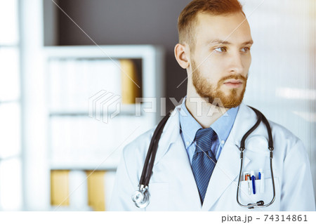 Red-bearded doctor standing straight in sunny clinic near his working place. Portrait of physician. Medicine concept Red-bearded doctor standing straight in sunny clinic near his working place. Portrait of physician. Medicine concept 74314861