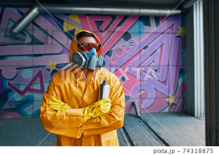 Graffiti painter in respirator mask standing near the wall with his paintings Graffiti painter in respirator mask standing near the wall with his paintings 74318875