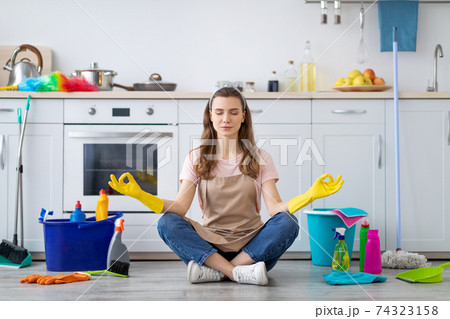 Young housewife surrounded by cleaning supplies meditating on floor at kitchen, taking break from house chores 74323158