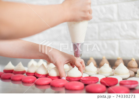 Step by step process of making macarons in confectionery shop. Close up horizontal shot of macarons shells in rows and hands of female pastry chef, filling it with white cream ganache 74330273