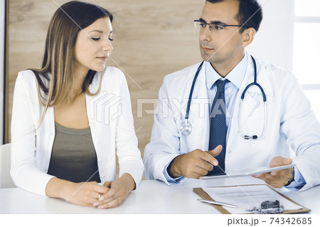 Doctor and patient discussing medical exam results while sitting at the desk in clinic. Male physician using tablet computer for filling up medication history record of young woman. Data in medicine Doctor and patient discussing medical exam results while sitting at the desk in clinic. Male physician using tablet computer for filling up medication history record of young woman. Data in medicine 74342685