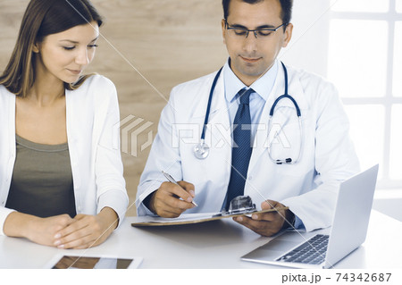 Doctor and patient discussing medical exam results while sitting at the desk in clinic. Male physician using clipboard for filling up medication history record of young woman. Data in medicine 74342687