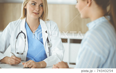 Woman-doctor and patient sitting and talking in hospital or clinic. Blonde therapist is cheerfully smiling. Medicine concept Woman-doctor and patient sitting and talking in hospital or clinic. Blonde therapist is cheerfully smiling. Medicine concept 74343254