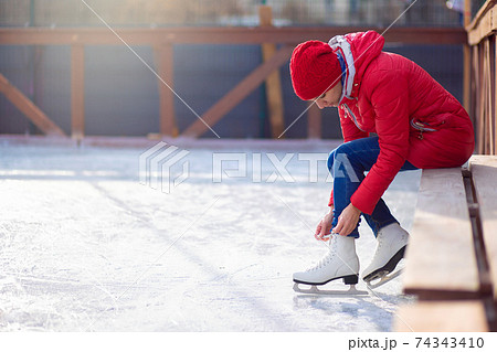 A girl in a red jacket sits on a bench on an open ice rink and laces figure skates. Winter sport A girl in a red jacket sits on a bench on an open ice rink and laces figure skates. Winter sport 74343410