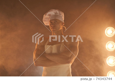 Portrait of muscular chef wearing white apron and chef hat, posing with folded arms on smoky background Portrait of muscular chef wearing white apron and chef hat, posing with folded arms on smoky background 74343744