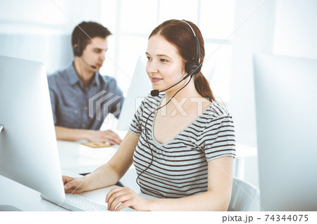 Casual dressed young woman using headset and computer while talking with customers online. Group of operators at work. Call center, business concept Casual dressed young woman using headset and computer while talking with customers online. Group of operators at work. Call center, business concept 74344075