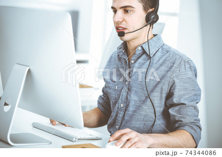 Casual dressed young man using headset and computer while talking with customers online. Call center, business concept Casual dressed young man using headset and computer while talking with customers online. Call center, business concept 74344086