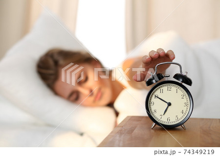 Sleepy young woman stretching hand to ringing alarm willing turn it off. Early wake up, not getting enough sleep, getting work concept. Sleepy young woman stretching hand to ringing alarm willing turn it off. Early wake up, not getting enough sleep, getting work concept. 74349219