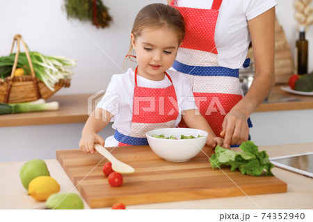 Happy family in the kitchen. Mother and child daughter cooking tasty breakfest of fresh salad. Little helper slicing and mix tomatos and greenery Happy family in the kitchen. Mother and child daughter cooking tasty breakfest of fresh salad. Little helper slicing and mix tomatos and greenery 74352940