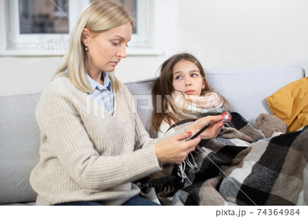 Sick child. Cute teen girl, lying on the sofa at home with scarf around neck, covered with blanket and looking at her mom, checking the temperature and calling to the doctor. Focus on sick girl Sick child. Cute teen girl, lying on the sofa at home with scarf around neck, covered with blanket and looking at her mom, checking the temperature and calling to the doctor. Focus on sick girl 74364984