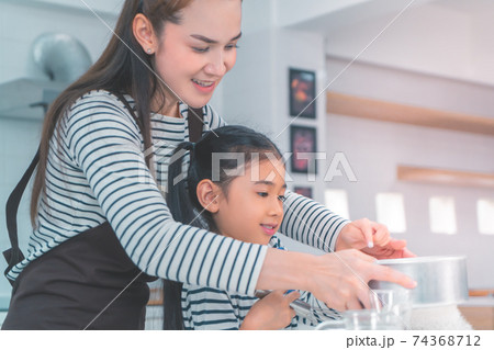 Little Asian girl is curiously watching her mother making bread and flour in home kitchen. Little Asian girl is curiously watching her mother making bread and flour in home kitchen. 74368712