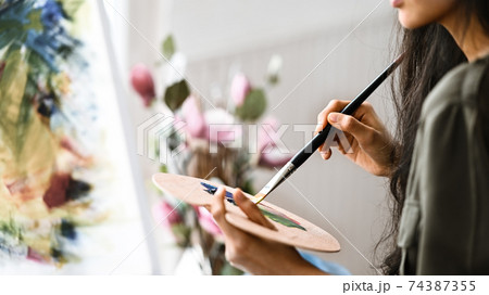 Cropped image of young beautiful artist girl sitting in front the drawing canvas while painting an oil color at the modern studio. Beautiful artist and gallery studio concept. Cropped image of young beautiful artist girl sitting in front the drawing canvas while painting an oil color at the modern studio. Beautiful artist and gallery studio concept. 74387355