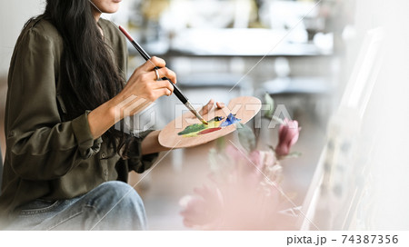 Cropped image of young beautiful artist girl sitting in front the drawing canvas while painting an oil color at the modern studio. Beautiful artist and gallery studio concept. Cropped image of young beautiful artist girl sitting in front the drawing canvas while painting an oil color at the modern studio. Beautiful artist and gallery studio concept. 74387356