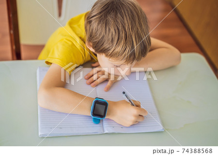 Little boy sitting at the table and looking smart watch. Smart watch for baby safety. The child makes school lessons, listening to music, calling friends Little boy sitting at the table and looking smart watch. Smart watch for baby safety. The child makes school lessons, listening to music, calling friends 74388568