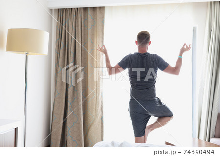 Man stands in lotus position by window Man stands in lotus position by window 74390494