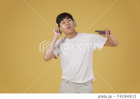 portrait of happy joyful cheerful young asian man wearing headphones listening to music on smartphone and dancing isolated on yellow background portrait of happy joyful cheerful young asian man wearing headphones listening to music on smartphone and dancing isolated on yellow background 74394915