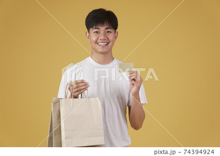 portrait of happy young asian man dressed casually holding shopping bags and credit card isolated on yellow background 74394924