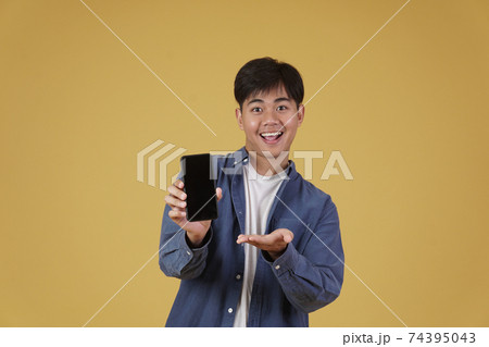 portrait of happy young asian man dressed casually smiling showing smartphone blank white screen isolated on yellow  background 74395043