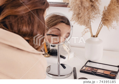 Young woman applying make-up to her face, looking at the round mirror 74395185
