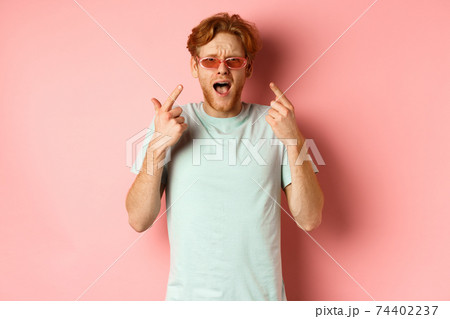 Young handsome man with red hair and beard pointing fingers at sunglasses, showing new glasses, standing over pink background 74402237